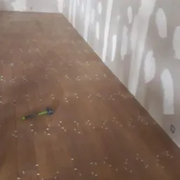 Układanie drewnopodobnych płytek podłogowych w pomieszczeniu z surowymi ścianami, widoczny młotek i pudełko na akcesoria do układania płytek.