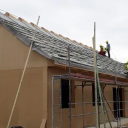 Budowa domu z drewnianych paneli: ekipa w kaskach i kamizelkach odblaskowych montuje dach na rusztowaniu, widoczne deski, membrana i drewniane elementy konstrukcyjne.