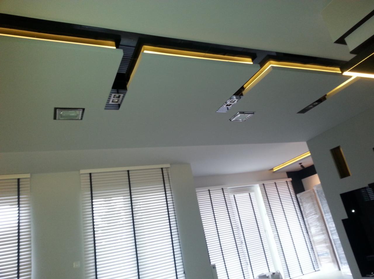 Nowoczesny sufit z podwieszanymi panelami i oświetleniem LED, widok z dołu na jasne wnętrze z żaluzjami.