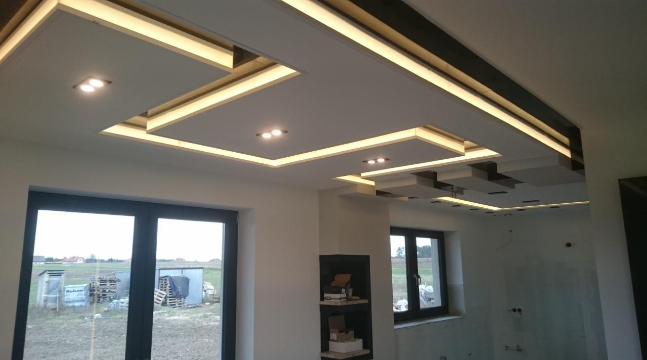 Nowoczesny sufit wielopoziomowy z podświetleniem LED i punktowymi lampami, widok na okno z krajobrazem wiejskim w tle, w pomieszczeniu w trakcie wykańczania.