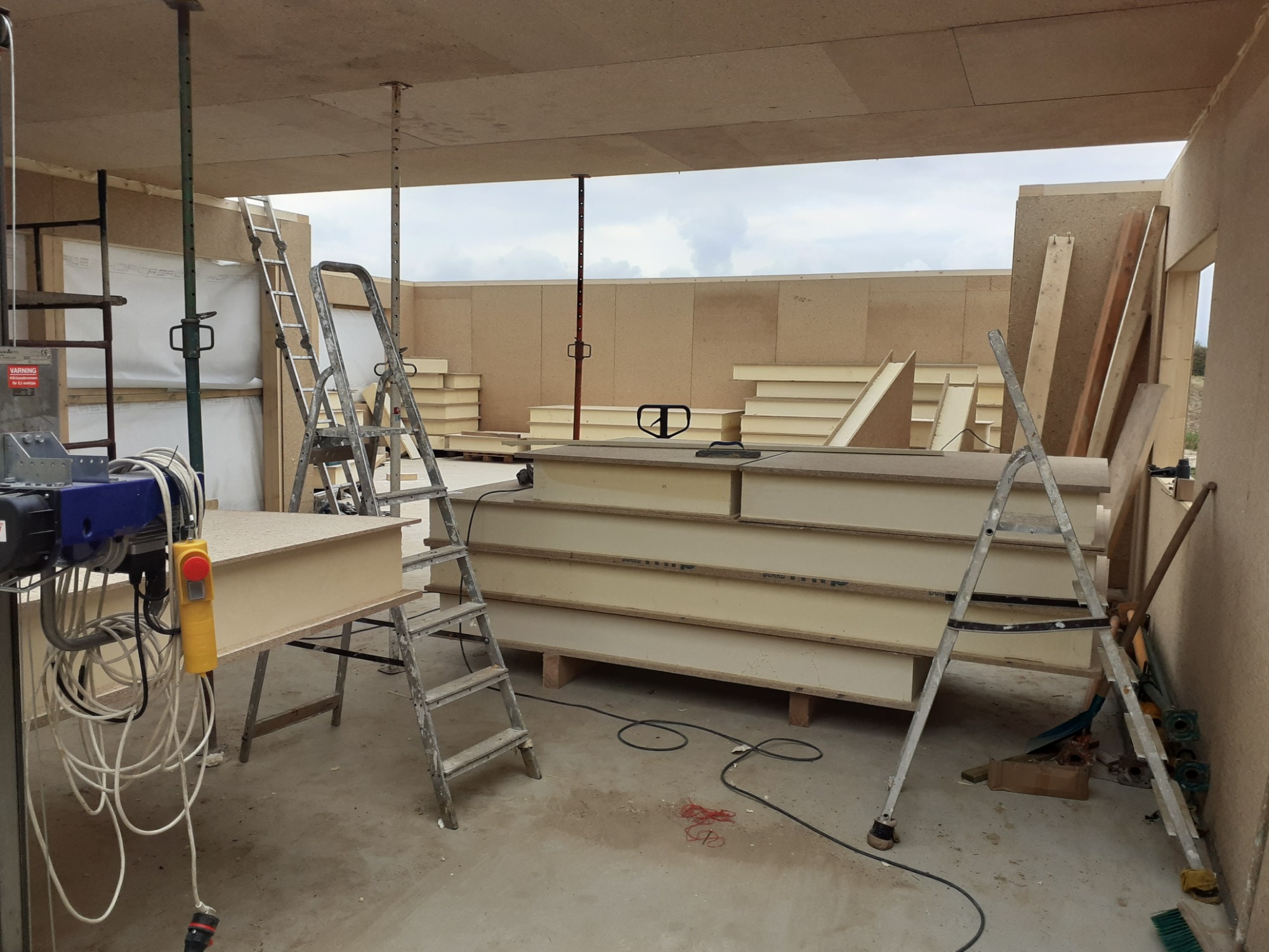 Wnętrze w trakcie budowy z drewnianych paneli, widoczne drabiny, podnośnik elektryczny z żółtym przyciskiem bezpieczeństwa oraz elementy konstrukcyjne w jasnym odcieniu drewna.