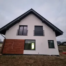 Nowoczesny dom z jasną elewacją i drewnianym panelem, czarnym dachem i oknami. Widoczne elementy wykończeniowe i otoczenie budynku.