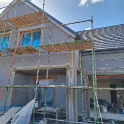 Dom w trakcie ocieplania z widocznymi rusztowaniami i szarymi płytami izolacyjnymi. Okna zabezpieczone folią. Fragment dachu z ciemnej dachówki.