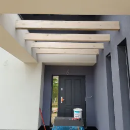 Wejście do domu z szarą elewacją i drewnianymi belkami pod zadaszeniem, widoczna puszka z farbą i narzędzia malarskie przy drzwiach.