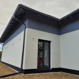 Elewacja domu po tynkowaniu, z widocznymi elementami orynnowania i oknem z materiałami izolacyjnymi wewnątrz.