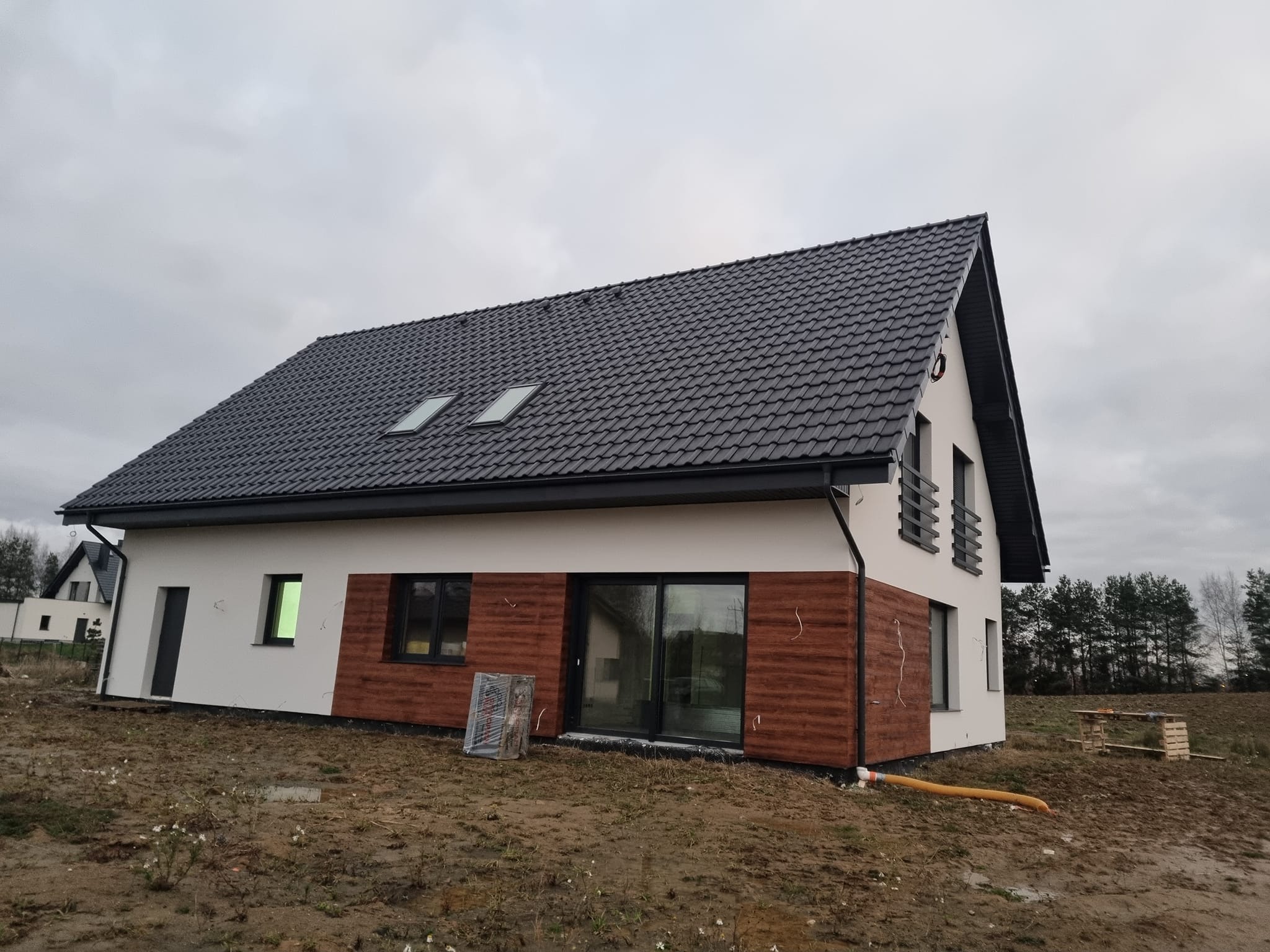 Nowoczesny dom jednorodzinny z dachem z dachówki ceramicznej i elewacją w kolorze białym z drewnianą okładziną. Widoczny fragment ogrodu i pochmurne niebo.