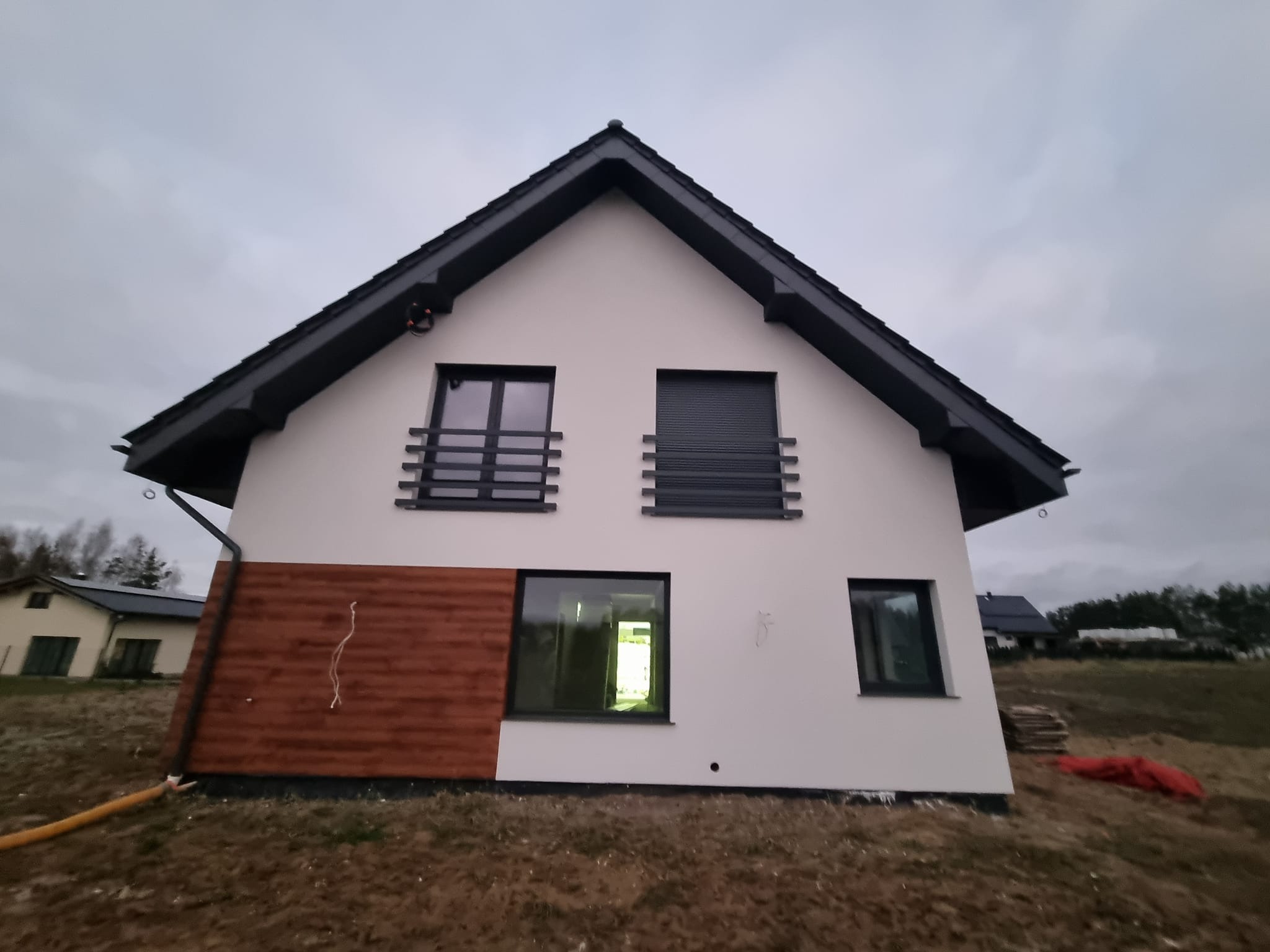 Nowoczesny dom z jasną elewacją i drewnianym panelem, czarnym dachem i oknami. Widoczne elementy wykończeniowe i otoczenie budynku.