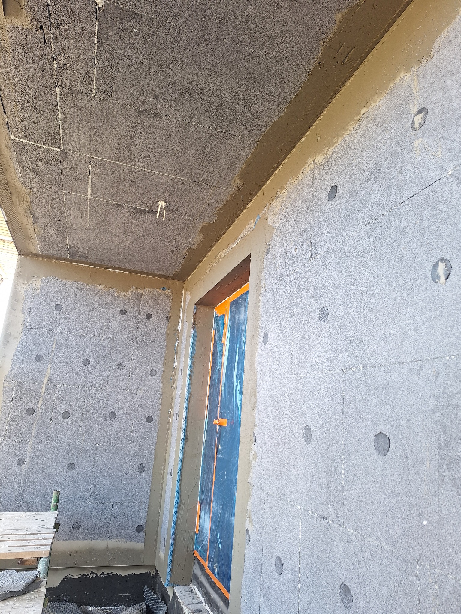 Ocieplenie ściany budynku styropianem grafitowym, widoczne kołki mocujące. Okno zabezpieczone folią. Prace w toku, widoczny fragment rusztowania.