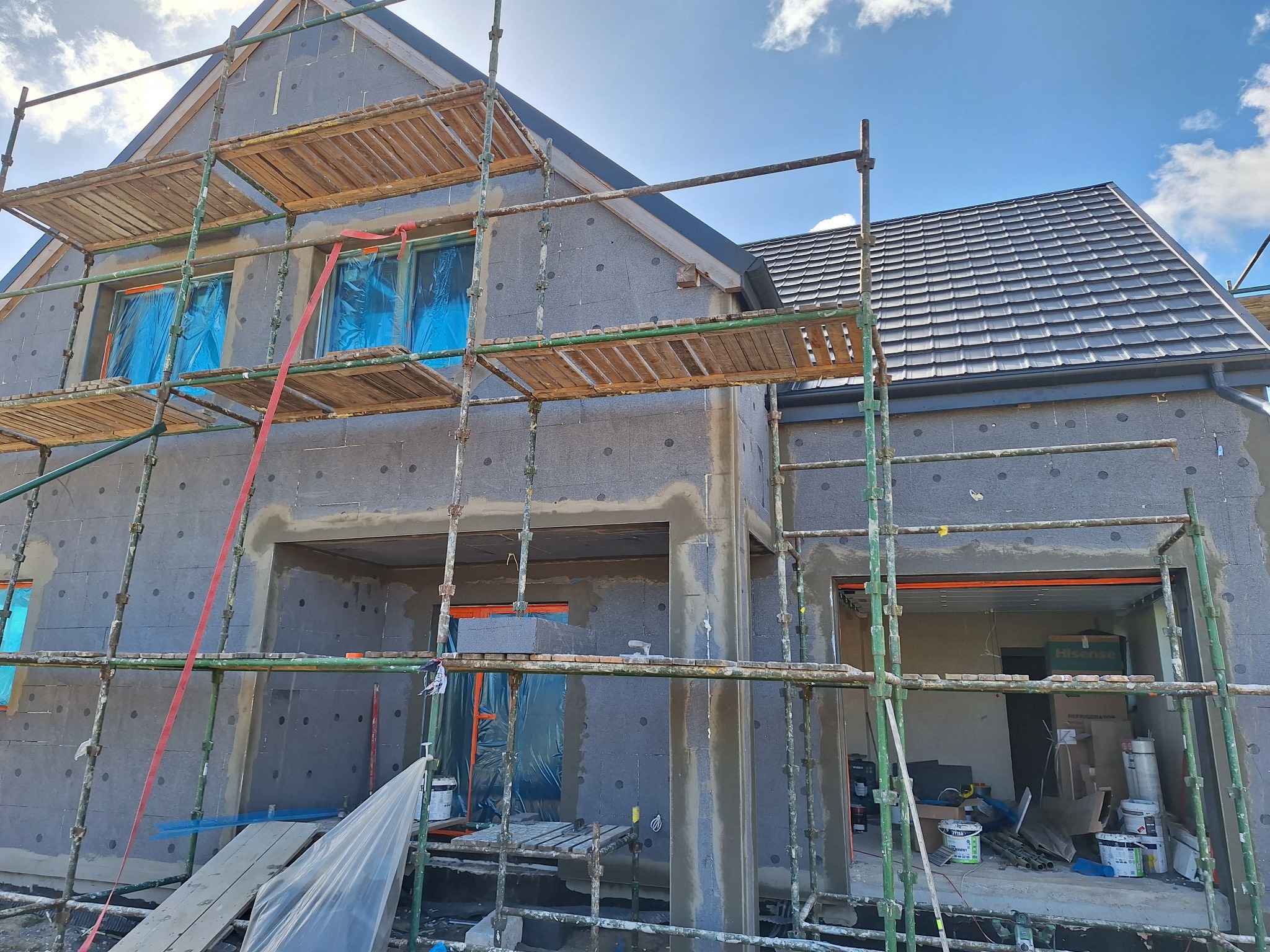 Dom w trakcie ocieplania z widocznymi rusztowaniami i szarymi płytami izolacyjnymi. Okna zabezpieczone folią. Fragment dachu z ciemnej dachówki.