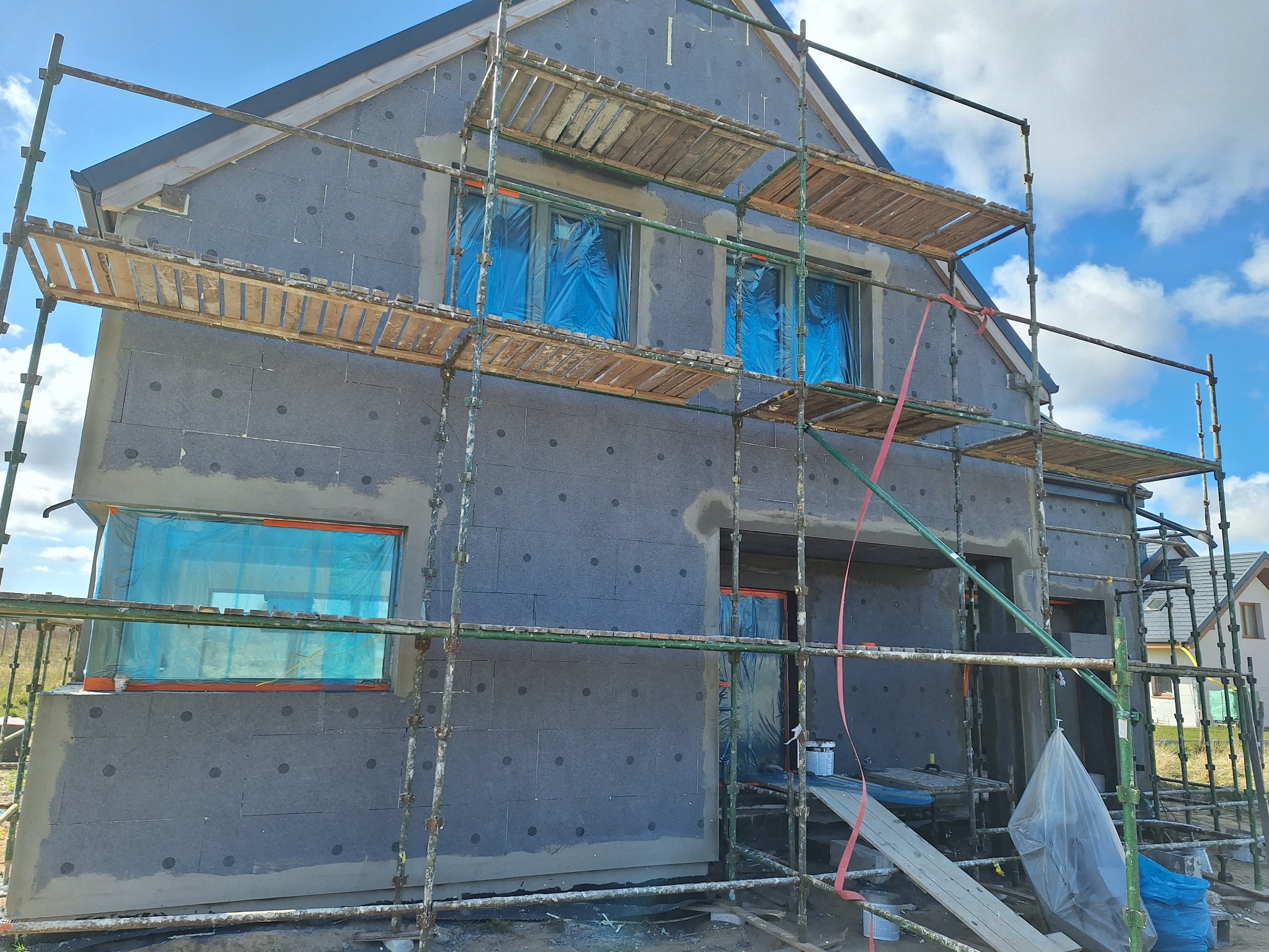 Dom w trakcie ocieplania, styropian z szarymi kołkami, rusztowanie, okna zabezpieczone folią, niebieskie niebo z chmurami w tle.