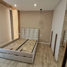 Beżowe łóżko tapicerowane z drewnianym stelażem i pikowanym zagłówkiem, obok biała szafa wnękowa z drewnianymi uchwytami, drewniana podłoga i dekoracyjne pionowe drewniane panele na ścianie.