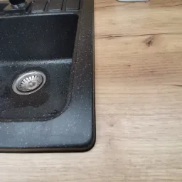 Czarny, nakrapiany zlew kuchenny z ociekaczem, zamontowany w blacie drewnopodobnym, z widocznym dozownikiem na płyn do naczyń.