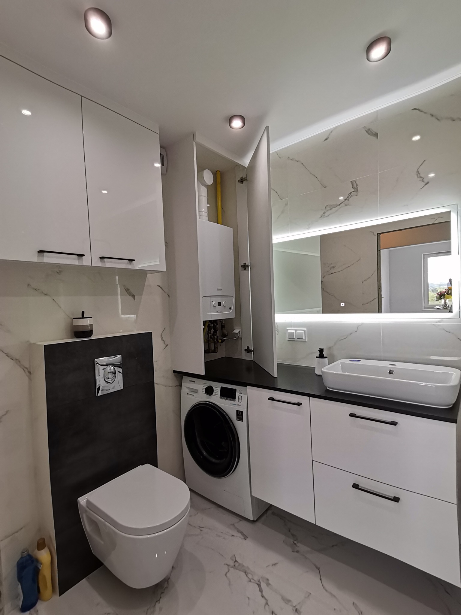 Nowoczesna łazienka z białymi meblami na wymiar, czarnym blatem, podświetlanym lustrem, marmurowymi płytkami i pralką ukrytą obok szafki, z wiszącą toaletą.