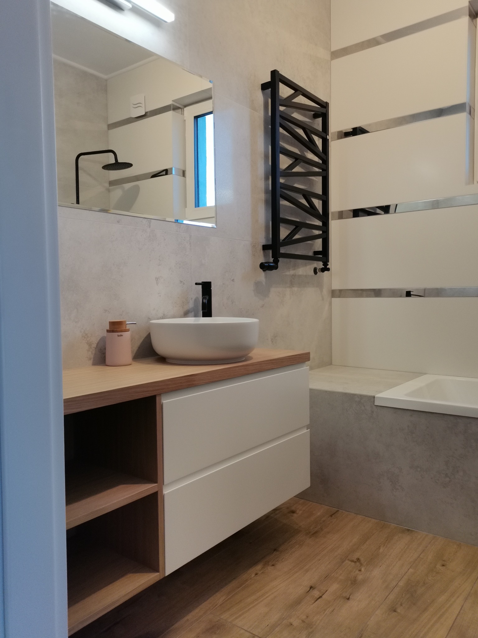 Nowoczesna łazienka z drewnianą podłogą, szafką pod umywalką z drewnianym blatem i białymi frontami, okrągłą umywalką nablatową, czarną armaturą i grzejnikiem drabinkowym.