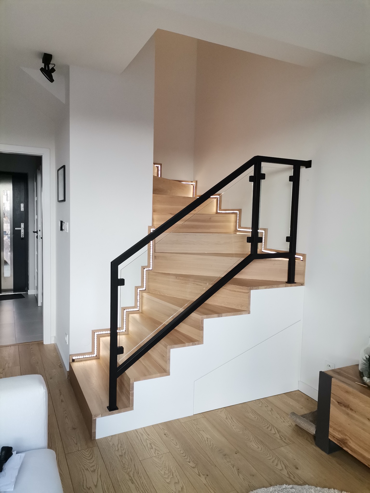 Nowoczesne schody drewniane z podświetleniem LED wzdłuż stopni, czarna balustrada z elementami szkła, białe ściany i drewniana podłoga.