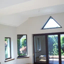 Jasne wnętrze z białymi ścianami, nietypowym trójkątnym oknem i dużymi przesuwnymi drzwiami balkonowymi z ciemnymi ramami, widok na zielony ogród.