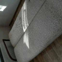 Jasnoszara kanapa z drewnianymi elementami w pokoju oświetlonym promieniami słońca padającymi na tapicerkę.