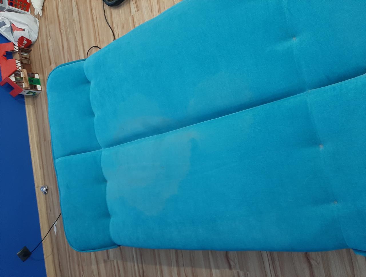 Turkusowa kanapa z widoczną plamą na siedzisku, sfotografowana z góry na tle drewnianej podłogi i niebieskiej ściany.