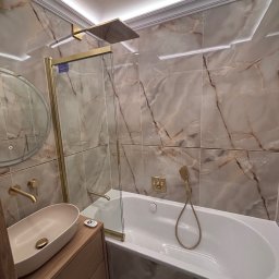 Zakład Ogólnobudowlany Degórski Leszek - Elegancka łazienka z beżowymi płytkami imitującymi marmur, złotą armaturą i podświetleniem LED. Umywalka nablatowa na drewnianej szafce, kabina prysznicowa ze złotymi ramami.
