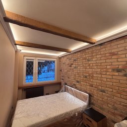 Zakład Ogólnobudowlany Degórski Leszek - Sypialnia z ceglaną ścianą, drewnianymi belkami pod sufitem i oknem. Łóżko przykryte folią ochronną. Oświetlenie LED podkreśla architekturę wnętrza.