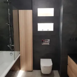 Nowoczesna łazienka z szarymi płytkami, podświetlaną wanną, wiszącą toaletą i drewnianymi akcentami, w tym wysoką szafką i szafką pod umywalkę.