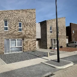 Nowoczesne, dwukondygnacyjne domy szeregowe z cegły klinkierowej w odcieniach brązu i szarości, z białymi ramami okiennymi i niewielkimi ogródkami z szarego grysu, otoczone brukowaną ulicą...