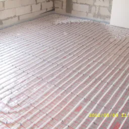 Rozłożona instalacja ogrzewania podłogowego w surowym stanie, widoczne białe rurki na czerwonej siatce, ściany w trakcie budowy.