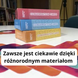 LinguaVision Edukacja & Języki - Stos trzech pudełek kart konwersacyjnych w kolorach różowym, niebieskim i żółtym, ułożonych na białym stole w klasie lub bibliotece.