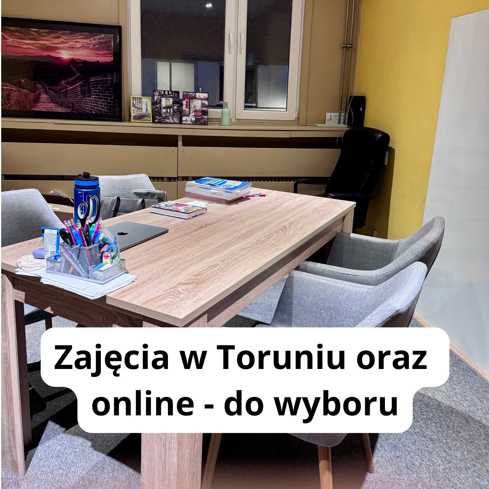 Stół w sali lekcyjnej z książkami, laptopem, przyborami do pisania i butelką wody. W tle widoczna tablica i krzesła.