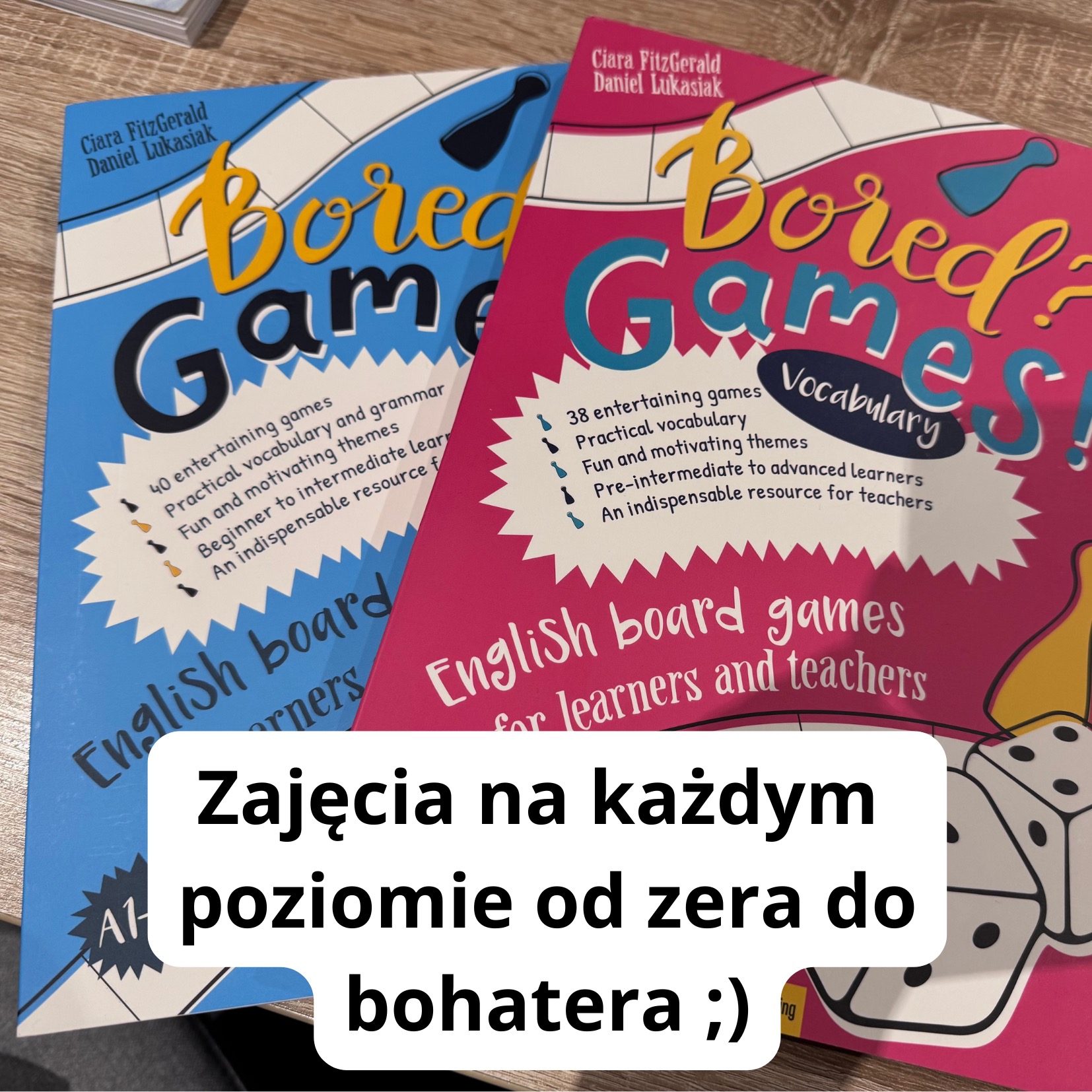 Dwa podręczniki do nauki języka angielskiego przez gry planszowe, jeden w kolorze niebieskim, drugi różowy, oba z tytułem 'Bored? Games!', na drewnianym stole.