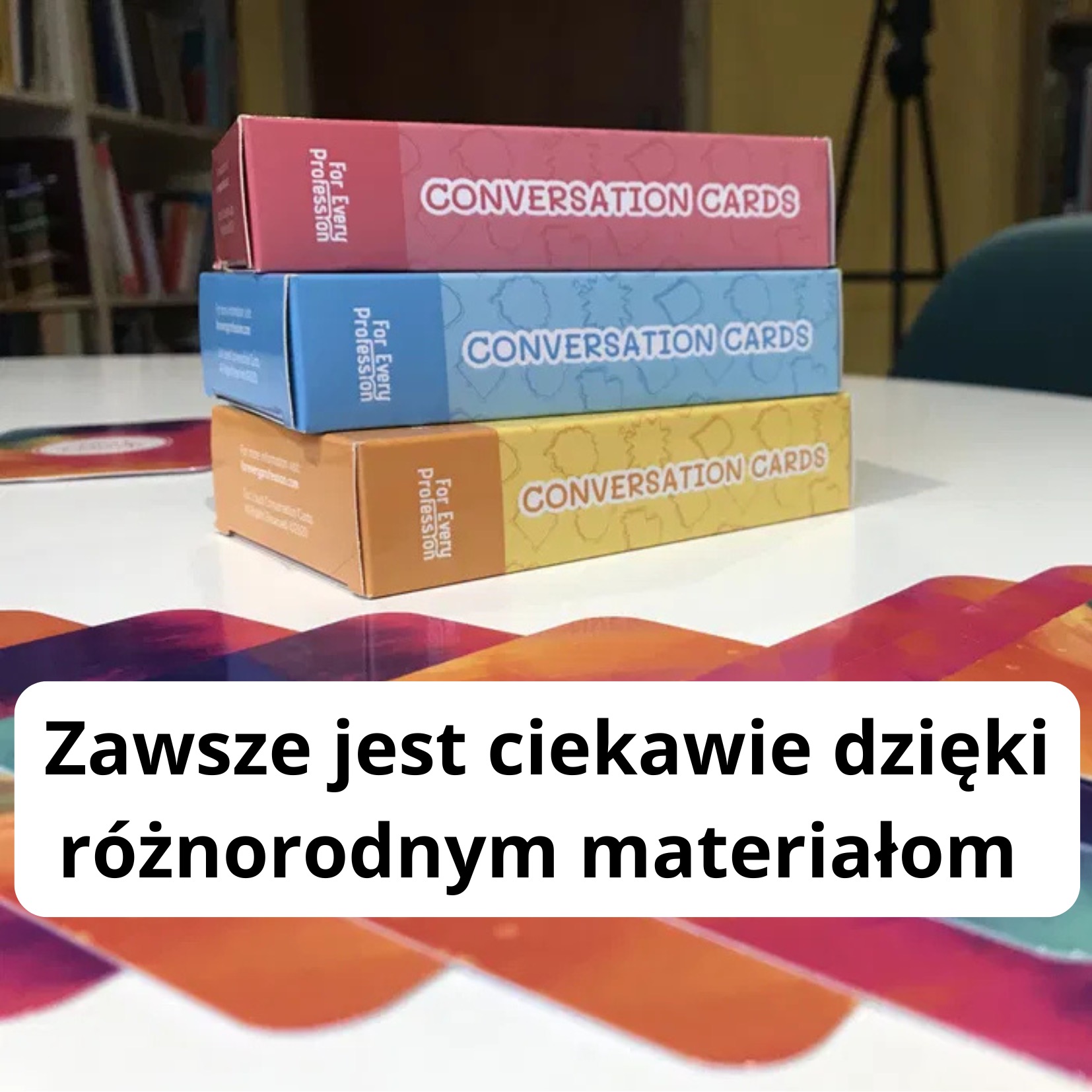 Stos trzech pudełek kart konwersacyjnych w kolorach różowym, niebieskim i żółtym, ułożonych na białym stole w klasie lub bibliotece.