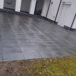 Szara kostka brukowa ułożona na podjeździe przed nowoczesnym domem z białymi ścianami i roletami zewnętrznymi, widoczne ślady wilgoci na powierzchni kostki.