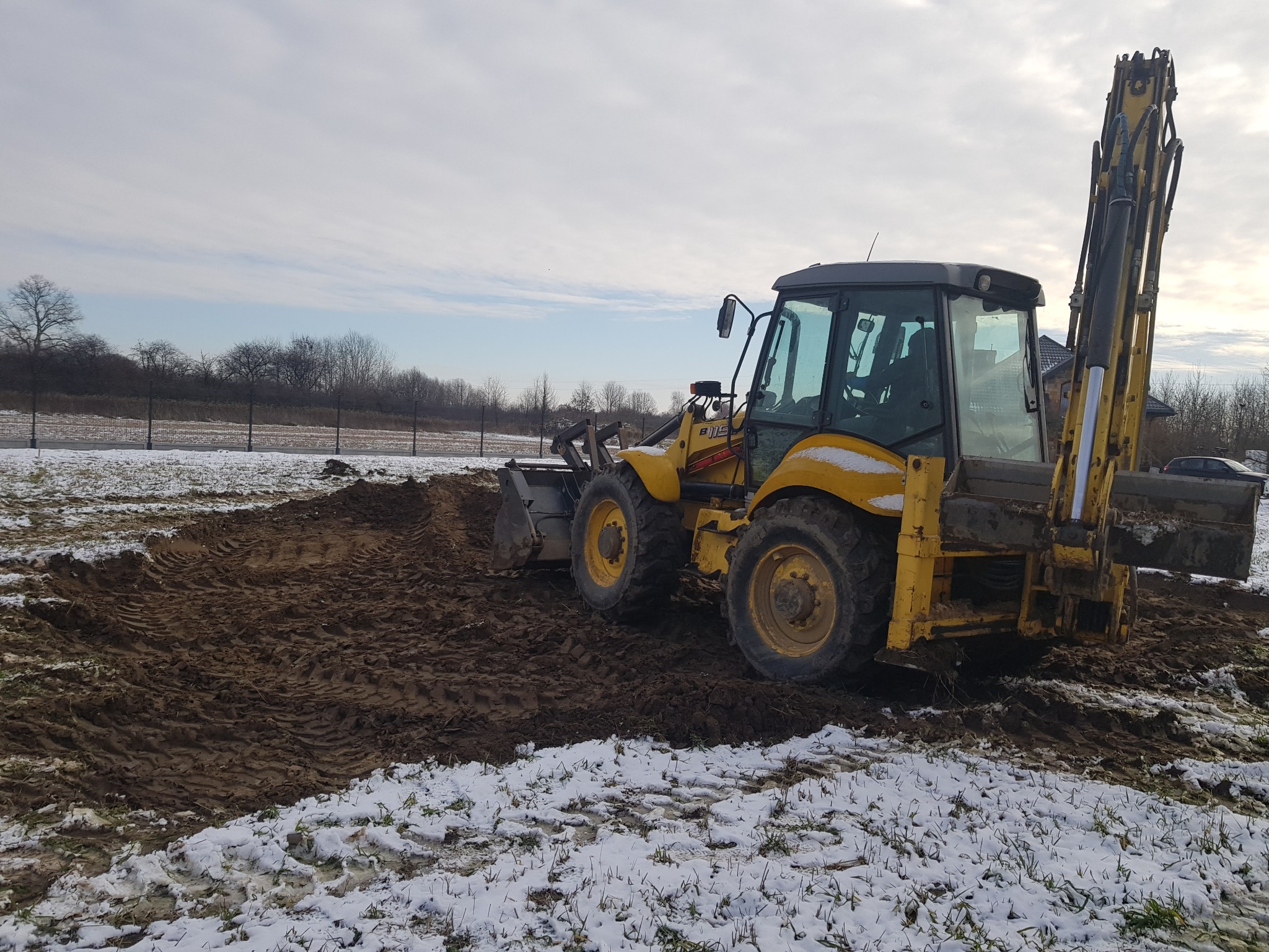 Żółta koparko-ładowarka przygotowuje teren pod trawnik na zasypanym śniegiem gruncie, widoczne ślady opon w błocie.
