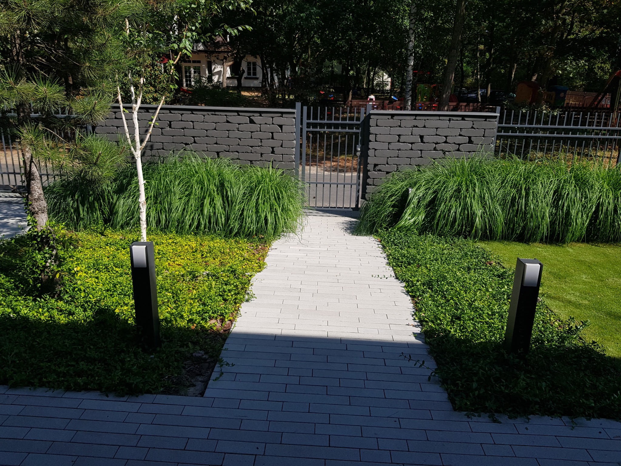 Ścieżka z jasnej kostki brukowej prowadząca przez zadbany ogród z ozdobnymi trawami i niską roślinnością, oświetlona lampami ogrodowymi, zakończona ciemnym murem z bramką.