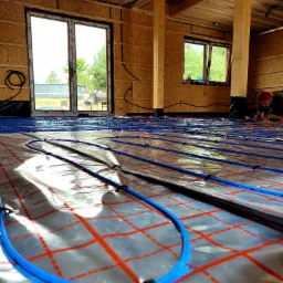 Instalacja ogrzewania podłogowego w surowym wnętrzu. Niebieskie rurki na folii z czerwoną kratką, widoczne okna i drewniana konstrukcja domu. Instalator w tle.