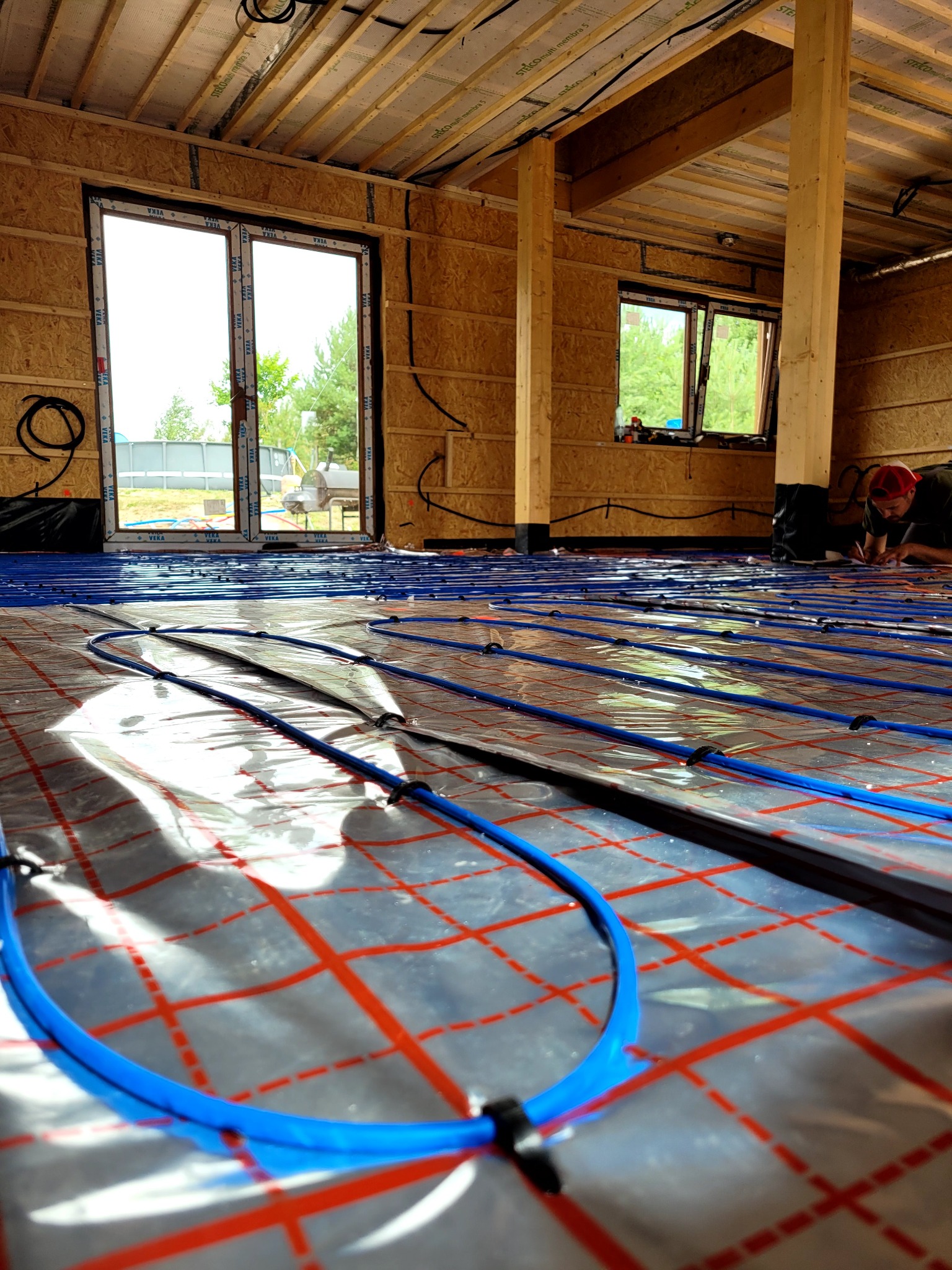 Instalacja ogrzewania podłogowego w surowym wnętrzu. Niebieskie rurki na folii z czerwoną kratką, widoczne okna i drewniana konstrukcja domu. Instalator w tle.