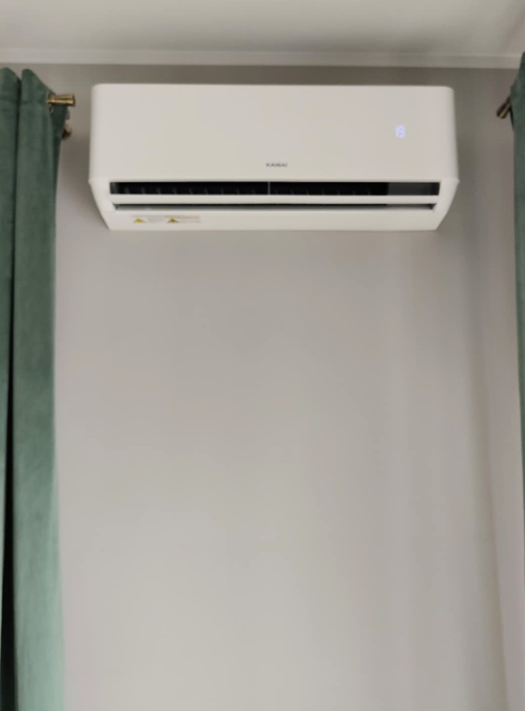 Biały klimatyzator marki Kaisai zamontowany na jasnej ścianie, z fragmentami zielonych zasłon po bokach kadru. Widoczny wyświetlacz z temperaturą 19 stopni.