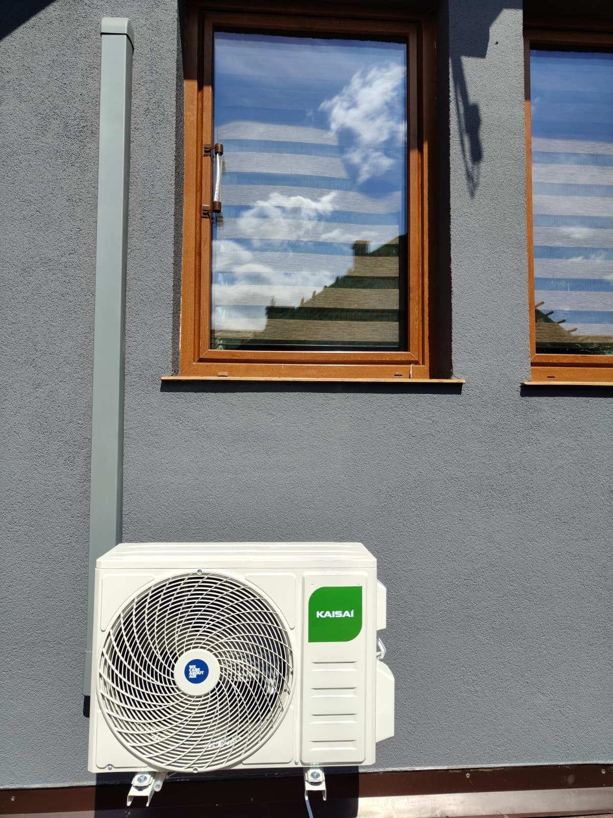 Zamontowana jednostka zewnętrzna klimatyzacji Kaisai na szarej elewacji budynku, widoczny fragment okna z odbiciem nieba. Ujęcie z dołu, słoneczny dzień.