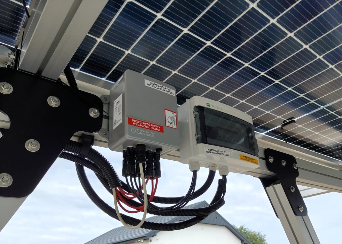 Rozdzielnica PV DC z wyłącznikiem przeciwpożarowym, element instalacji fotowoltaicznej zamontowany pod panelami słonecznymi. Widoczne przewody elektryczne i elementy mocujące.