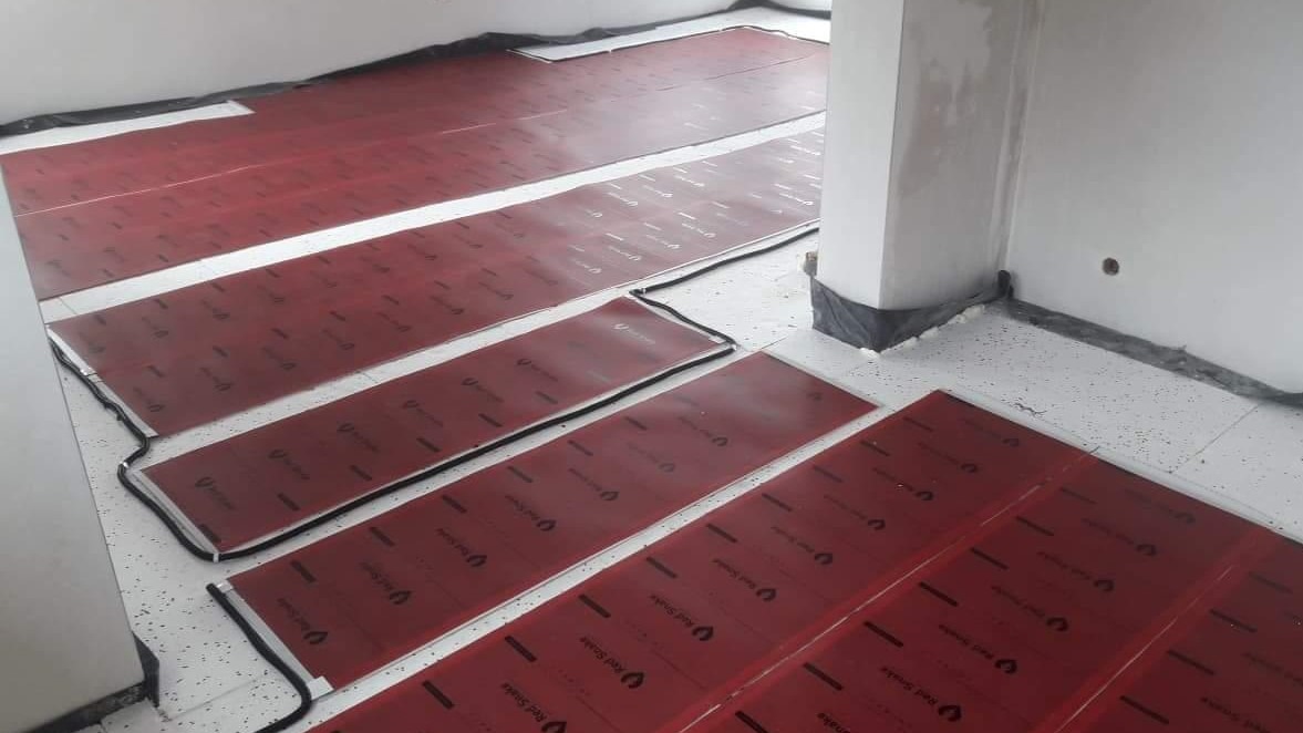 Czerwone maty grzewcze elektryczne marki Red Snake rozłożone na podłodze w trakcie instalacji, widoczne czarne przewody zasilające i fragment ściany z zabezpieczeniem folią.