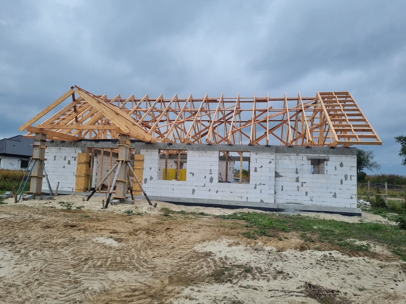 Budowa domu jednorodzinnego: ściany z bloczków, drewniana konstrukcja dachu w trakcie montażu, widoczne podpory i elementy szalunku.