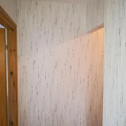 Korytarz z nową tapetą w pionowe, nieregularne paski w odcieniach szarości i srebra, widoczny fragment drewnianej futryny drzwi po lewej stronie.