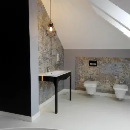 Nowoczesna łazienka na poddaszu ze skośnym sufitem, umywalką na czarnym postumencie z podświetleniem LED, toaletą i bidetem na tle ściany wyłożonej płytkami imitującymi kamień.