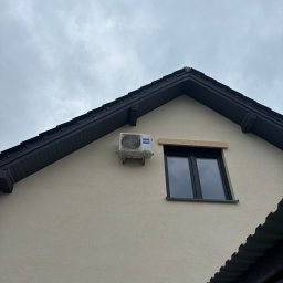 OPTIMAL-PLAST Rafał Pajdak - Klimatyzator zamontowany na elewacji domu z ciemną dachówką i oknem. Widok z dołu na szarym tle nieba.