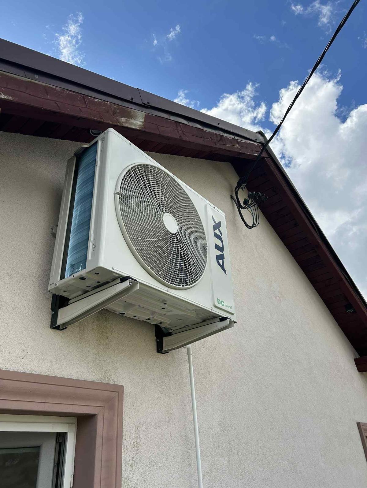 Biała jednostka zewnętrzna klimatyzacji AUX zamontowana na elewacji domu z brązowym dachem, widoczne przewody i uchwyty montażowe. Błękitne niebo w tle.