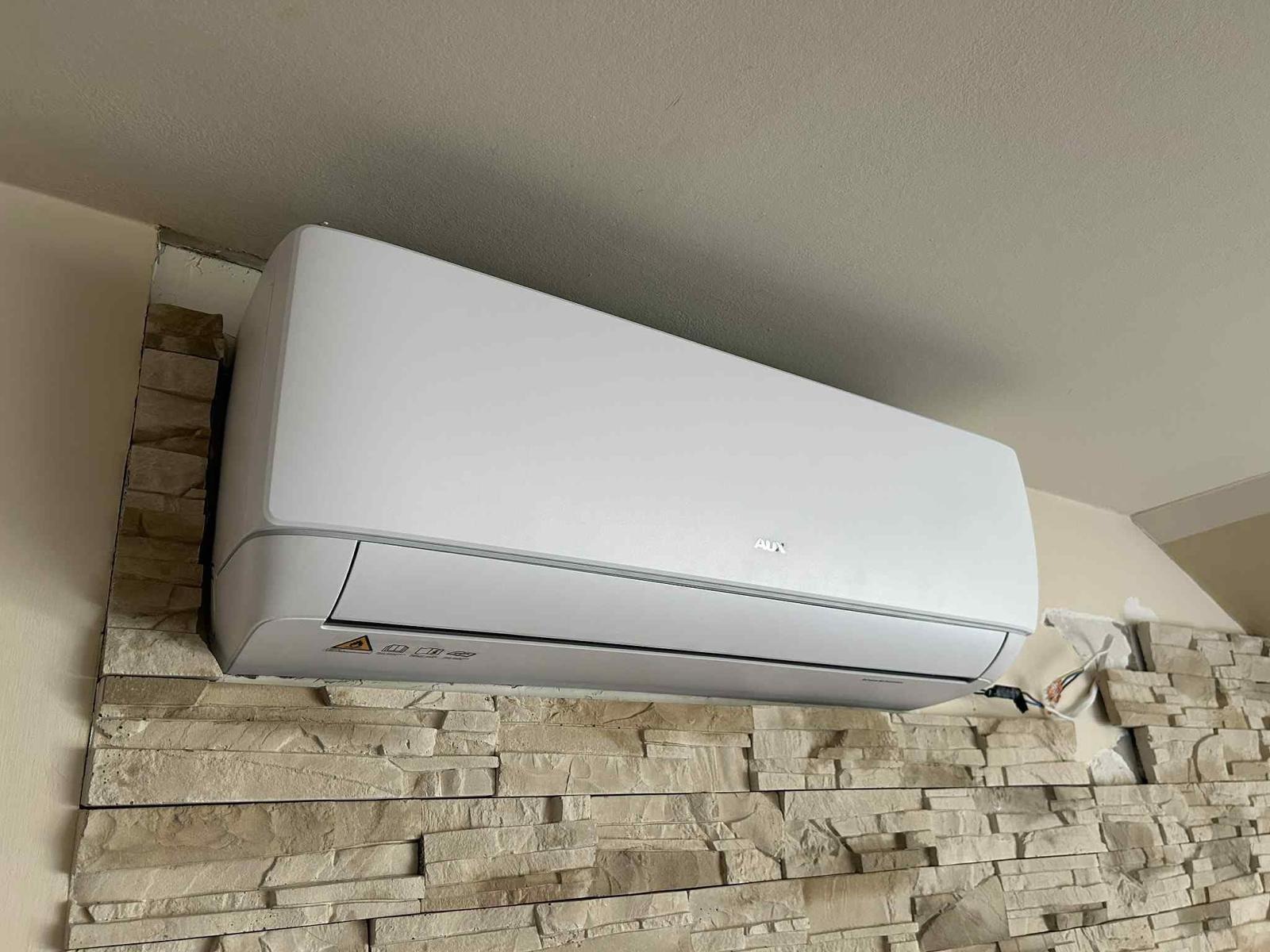 Biały klimatyzator AUX zamontowany na ścianie z dekoracyjnej cegły w jasnym odcieniu. Widoczne przewody elektryczne. Ujęcie z dołu, lekki kąt.