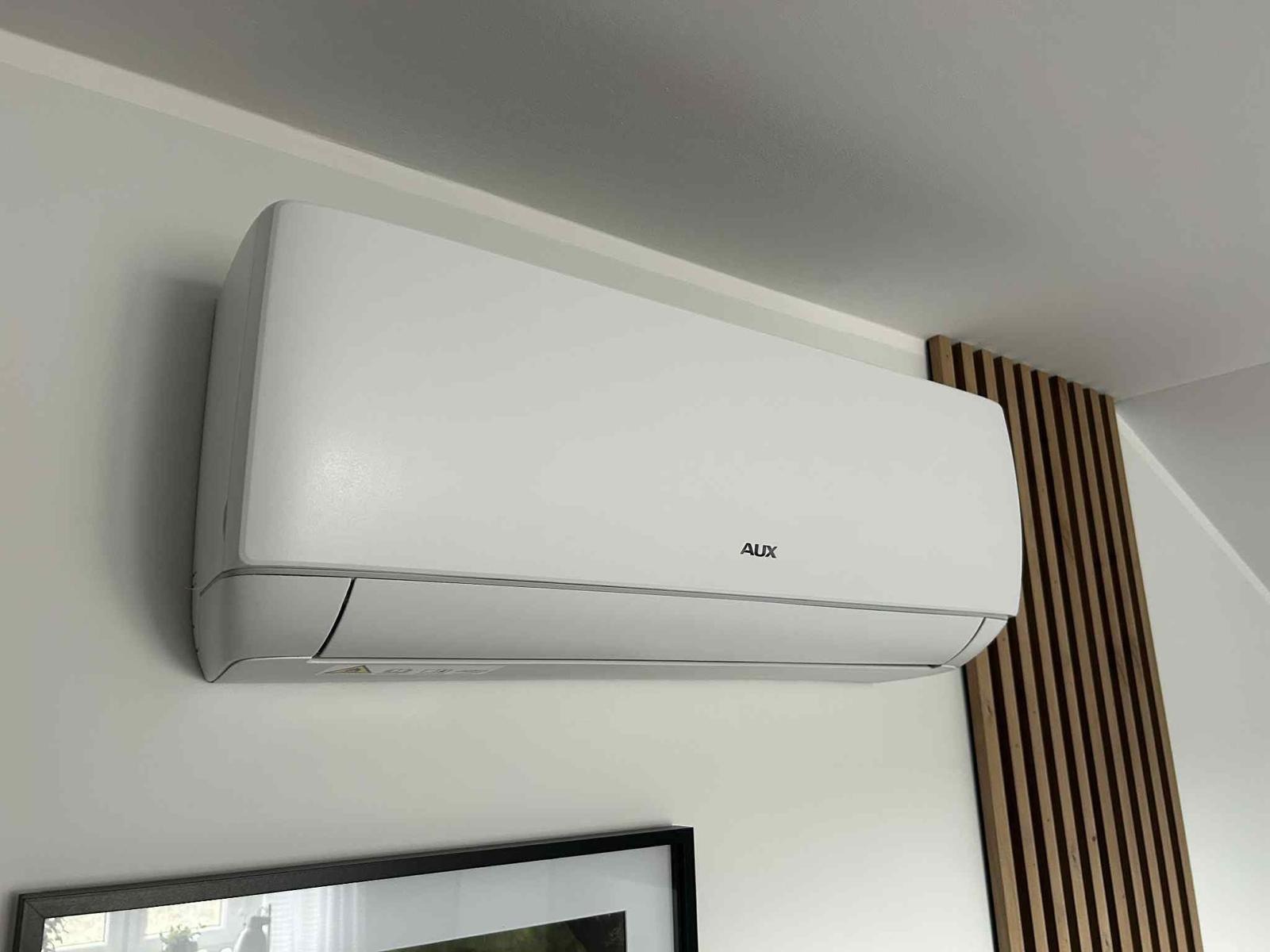 Biała klimatyzacja AUX na ścianie w jasnym wnętrzu, widoczny fragment dekoracyjnej drewnianej ścianki i ramy obrazu. Minimalistyczny design.