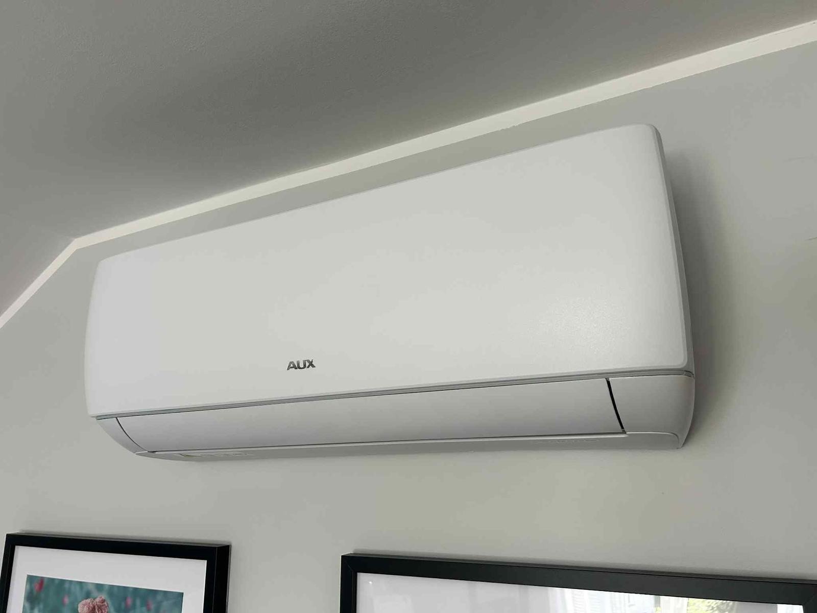 Biała klimatyzacja AUX zamontowana na szarej ścianie w pomieszczeniu. Pod spodem widoczne fragmenty dwóch obrazów w czarnych ramkach.