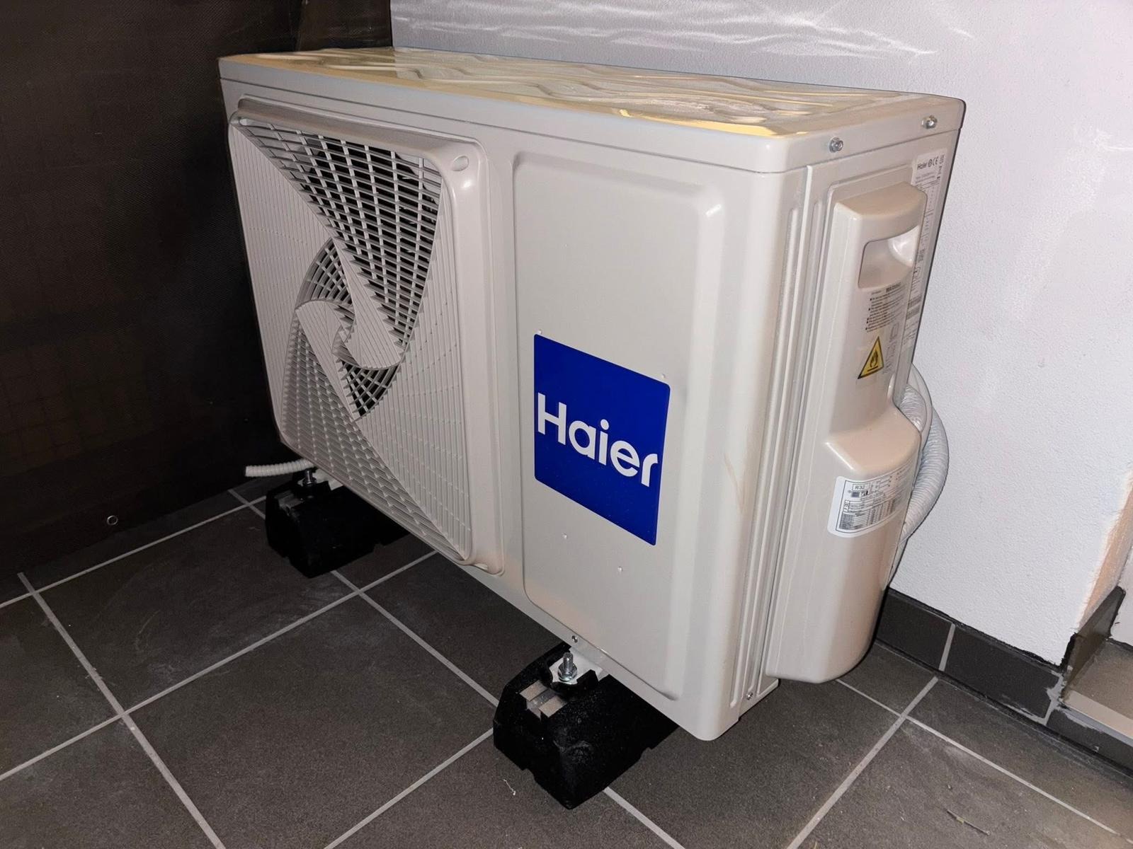 Biała jednostka zewnętrzna klimatyzacji Haier, zamontowana na czarnych podkładkach, na szarej podłodze z płytek, przy białej ścianie. Widok z boku.
