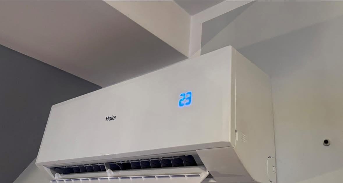 Biały klimatyzator Haier na ścianie, wyświetlający temperaturę 23 stopnie, zamontowany w rogu pomieszczenia z szarą ścianą.